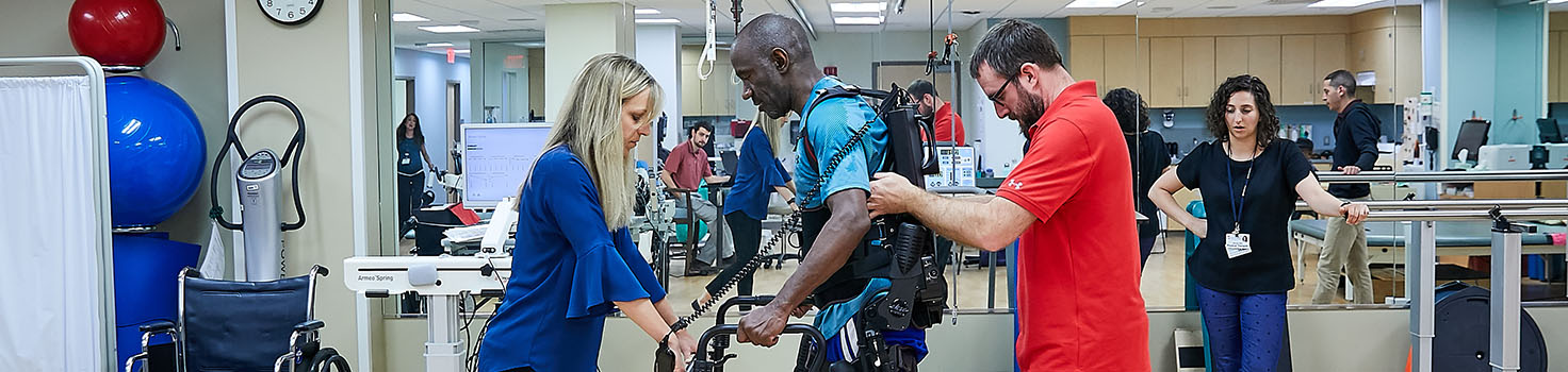 African American man walking in the EksoGT exoskeleton with the help of two physical therapists after suffering a spinal cord injury.