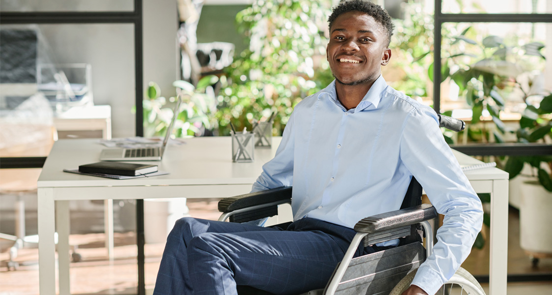 Portrait of African businessman with disability sitting in wheelchair and smiling at camera during his work at office