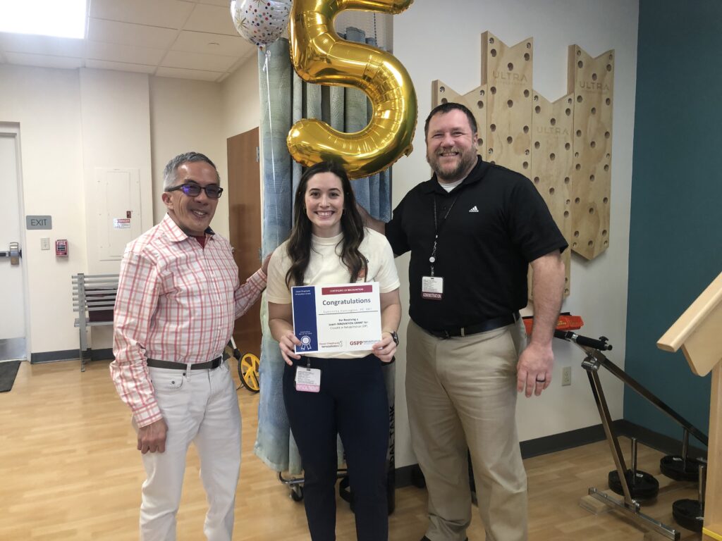 Three people smile for a photo. The person in the middle is holding a certificate that reads "Congratulations"