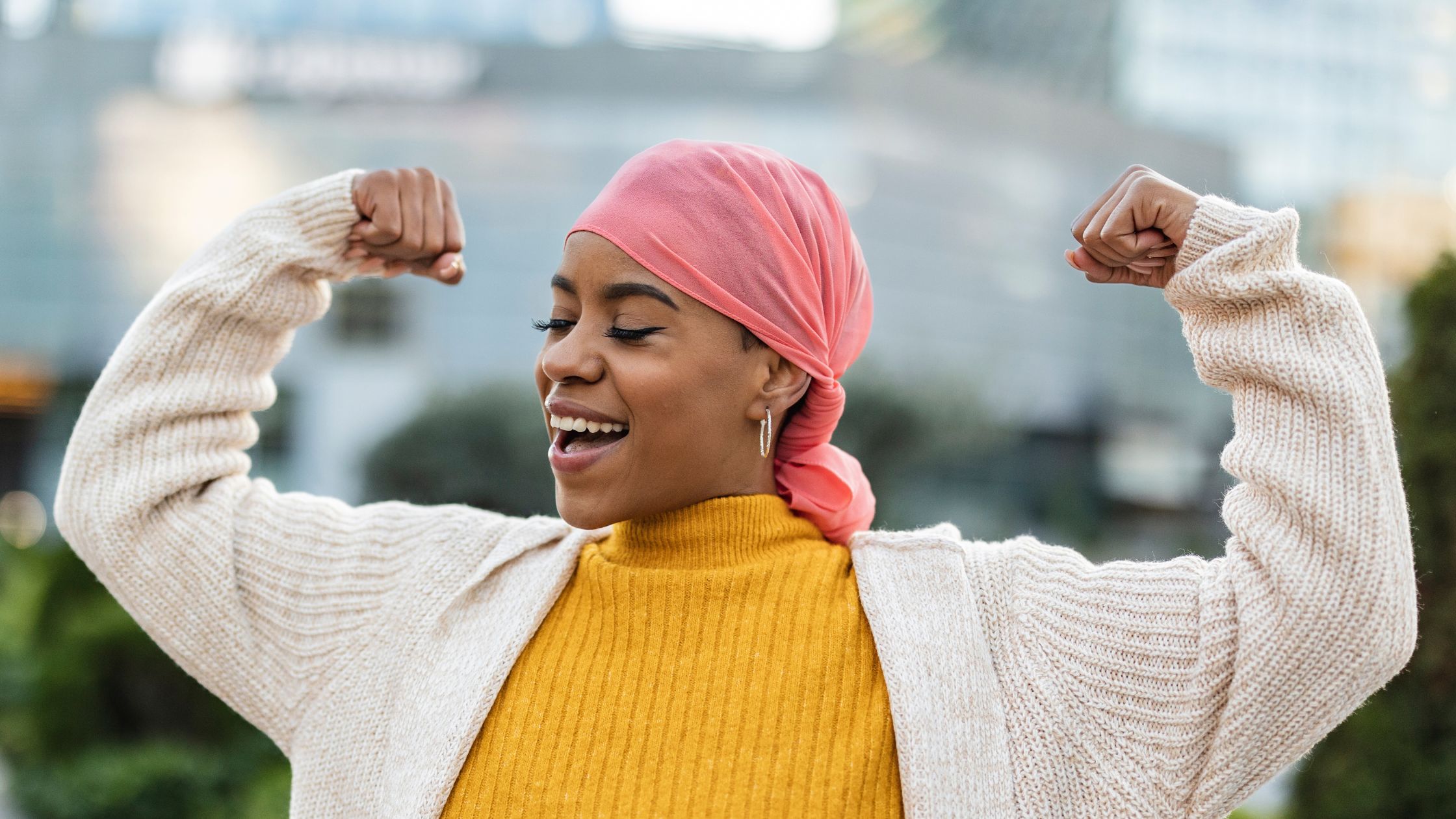 A person wearing a yellow top and tan sweater makes muscles and smiles while outside